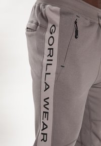 Šviesiai pilki sportiniai džinsai su siaurėjimu, turintys šonines kišenes, elastinį liemenį ir ryškų juodą "GORILLA WEAR" logotipą šone.