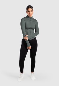 Chaqueta deportiva de color verde oscuro con cremallera, de manga larga y diseño ajustado, combinada con leggings negros. Modelo llevando zapatillas blancas y calcetines tipo crew.