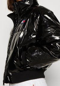 Osoba oblečená do lesklé černé puffer bundy s logem Tommy Jeans, se zipem a černými žebrovanými manžetami a spodním lemem.