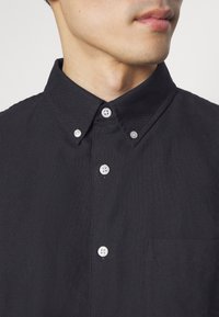 Camicia nera con bottoni a contrasto, dotata di colletto e taschino sul petto. Il tessuto appare texturizzato ed è tagliato per una vestibilità aderente.