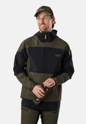 Mężczyzna w zielono-czarnej kurtce outdoorowej i czapce, trzyma czarną butelkę na wodę z pomarańczowymi sylwetkami zwierząt.