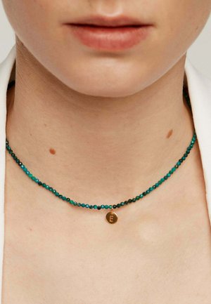Primer plano del cuello de una persona que lleva un collar de cuentas en color teal con un pequeño colgante de oro grabado con la letra E.