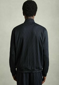 Veste noire texturée avec un motif en vagues, dotée de manches longues et d'un col, vue de dos sur un fond neutre.