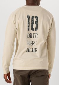 Beige Langarmshirt mit einem abgenutzten schwarzen Grafiktextdesign auf dem Rücken, das "18 BUTCHER OF BLUE" liest. Aus weichem Baumwollmaterial.