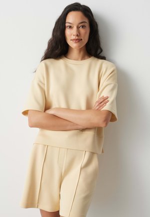 Frau mit langen dunklen Haaren, die ein beige kurzärmeliges Oberteil und passende Shorts trägt, steht mit verschränkten Armen an einer weißen Wand.