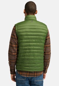 Grön pufferväst med hög krage, quiltat mönster och glänsande material, bärs över en brun rutig skjorta med blå och krämfärgade linjer.