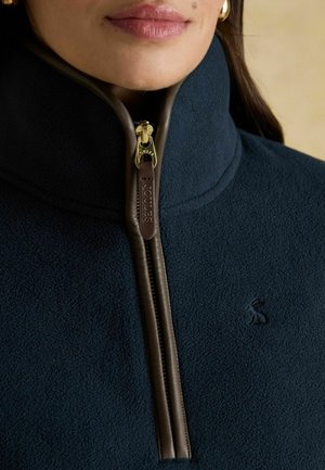 Woman wearing navy fleece jacket with brown leather-trimmed zipper and gold zipper pull, featuring subtle embroidered logo on chest.