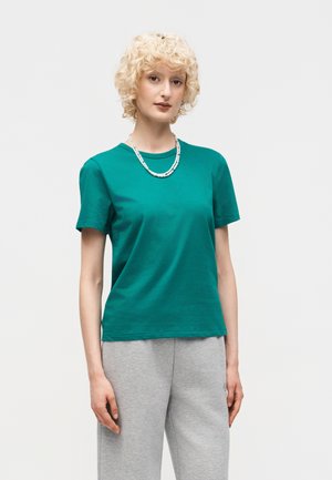 Młoda kobieta o kręconych blond włosach, ubrana w zielony t-shirt, szare spodnie dresowe i biały naszyjnik z koralików, stojąca na jednolitym tle.