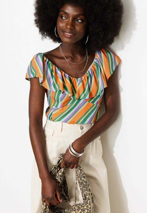 Femme aux grands cheveux bouclés portant un haut coloré à rayures et volants ainsi qu’un pantalon couleur crème, tenant un sac imprimé léopard avec des bracelets argentés.