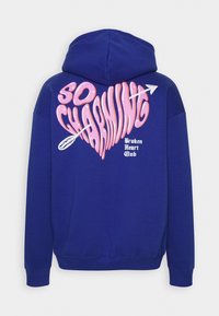 Královská modrá mikina s kapucí s velkou růžovou grafikou srdce s nápisem "SO CHARMING" a šipkou, pod ní pak "Broken Heart Club".