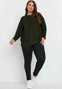 Pull en maille texturée vert foncé avec coupe ample, manches longues et col rond, associé à un jean skinny noir et des baskets blanches.