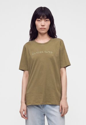 Kvinde iført en olivengrøn Michael Kors T-shirt med små pynt og lyseblå jeans, stående mod en ensfarvet hvid baggrund.