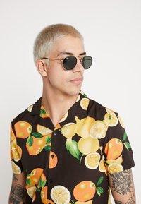 Short-sleeve shirt with a black background and a colorful fruit print featuring oranges and lemons. Model wears dark sunglasses.