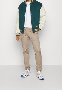 Grön och krämfärgad varsityjacka med knäppning, randiga ribbade muddar, tillsammans med beige byxor och vita sneakers med blå detaljer.