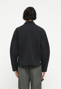 Casaco preto oversized com uma textura suave e uma bainha ligeiramente arredondada. Apresenta detalhes de costura discretos e um pequeno logotipo na parte inferior.