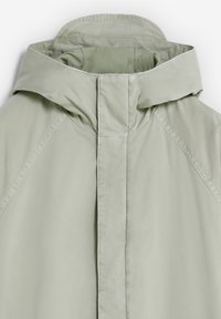 Vihreä takki hupulla, jossa on sileä pinta ja puhdas, minimalistinen muotoilu. Saumojen varrella näkyvät ompeleet, ei metalliosia tai koristeita.