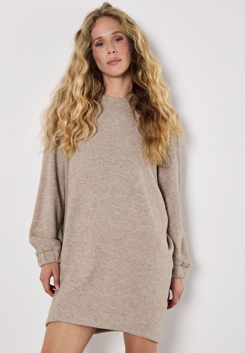 Beige trui-jurk met een gestructureerde afwerking, lange mouwen en een ronde halslijn, die tot halverwege de dij reikt. Naaddetails voegen visuele interesse toe.