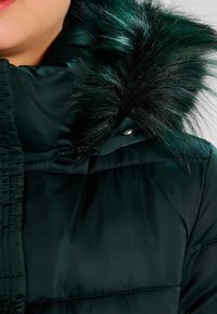 Giacca imbottita verde scuro con colletto alto e rifiniture in finta pelliccia nera. Presenta una chiusura con zip e bottoni nella zona del collo.