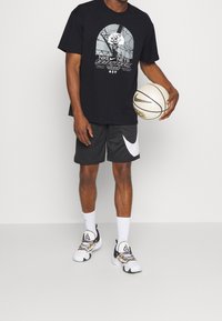 Camiseta gráfica negra con diseño de baloncesto en blanco y gris, pantalones cortos grises con logo blanco de Nike, calcetines blancos y zapatillas de baloncesto multicolores.