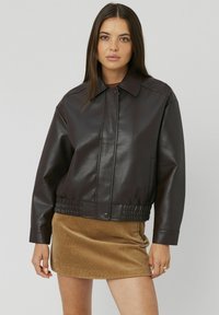 Blouson en cuir marron, avec un col classique, une fermeture éclair et une taille élastiquée. Porté par-dessus une jupe en velours côtelé beige.