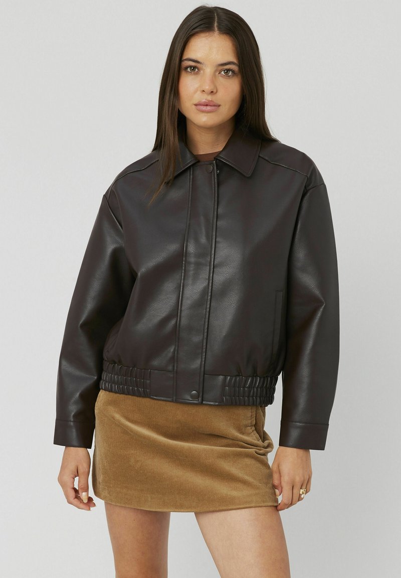 Blouson en cuir marron, avec un col classique, une fermeture éclair et une taille élastiquée. Porté par-dessus une jupe en velours côtelé beige.