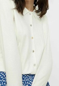 Chemise blanche boutonnée avec col Claudine, ornée de boutons variés et d'une texture douce, associée à une jupe bleue à motif.