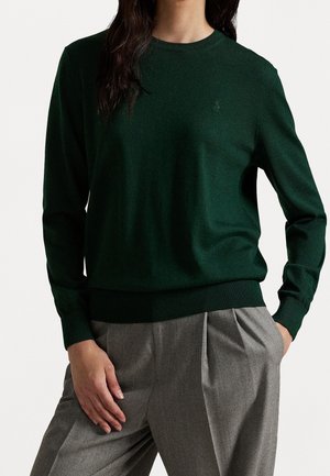 Femme portant un pull à manches longues de couleur vert foncé associé à un pantalon plissé gris taille haute, une main dans la poche, debout devant un fond uni.