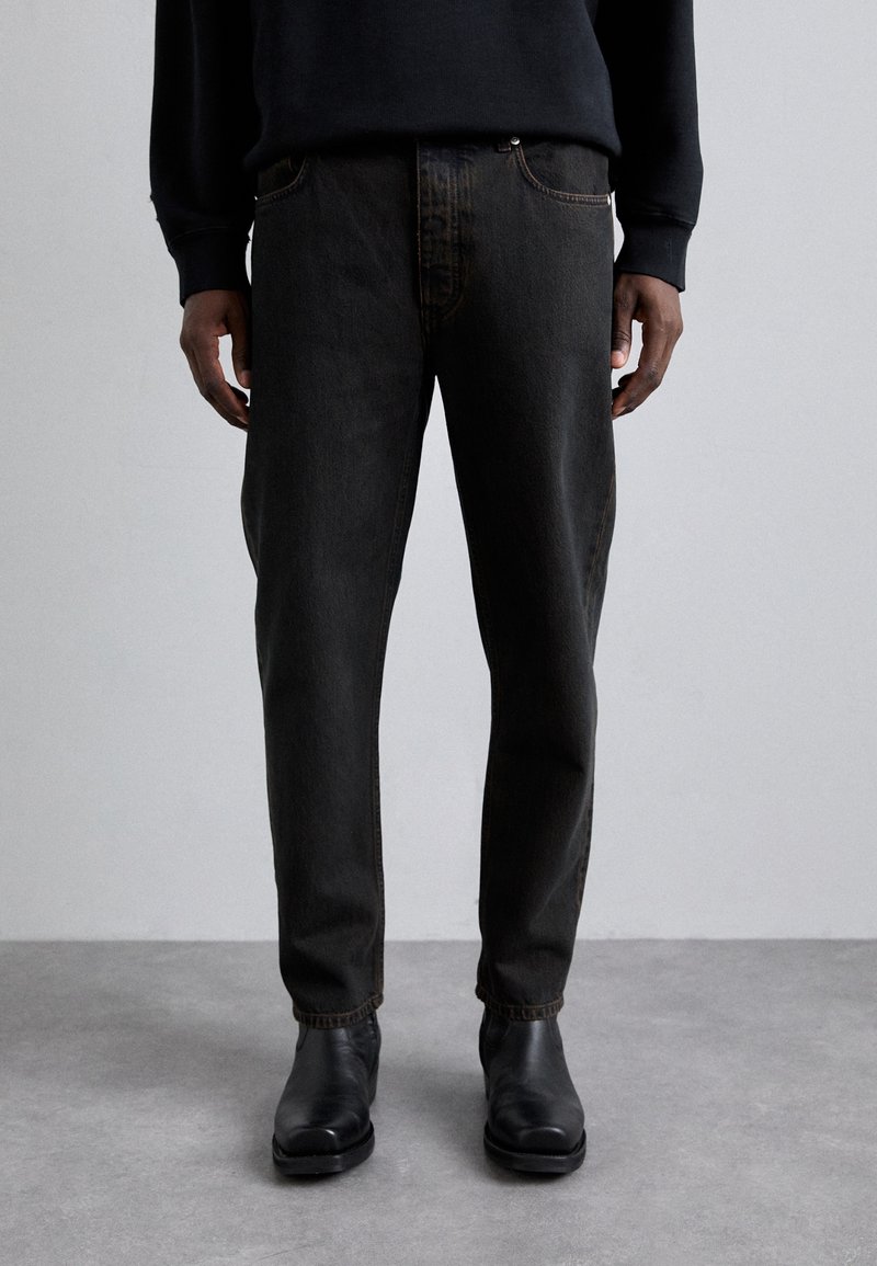 Mustadest denimist püksid sirge lõikega, kontrastse õmblusega ja eesmistest taskutest, koos mustade pahkluuni ulatuvate saapadega. Matte tekstuur.