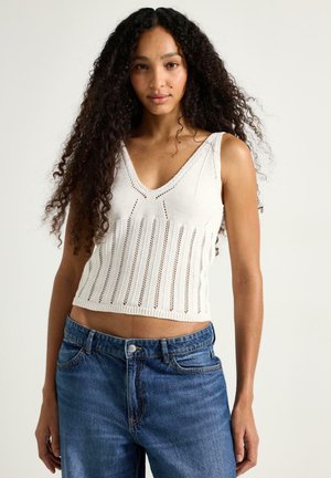Donna con lunghi capelli ricci che indossa un top bianco a maglia senza maniche con scollo a V e jeans di denim blu, davanti a uno sfondo semplice.