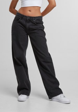 Kvinde iført løse sorte jeans og hvide sneakers, stående med hænderne bag ryggen mod en ensfarvet grå baggrund.