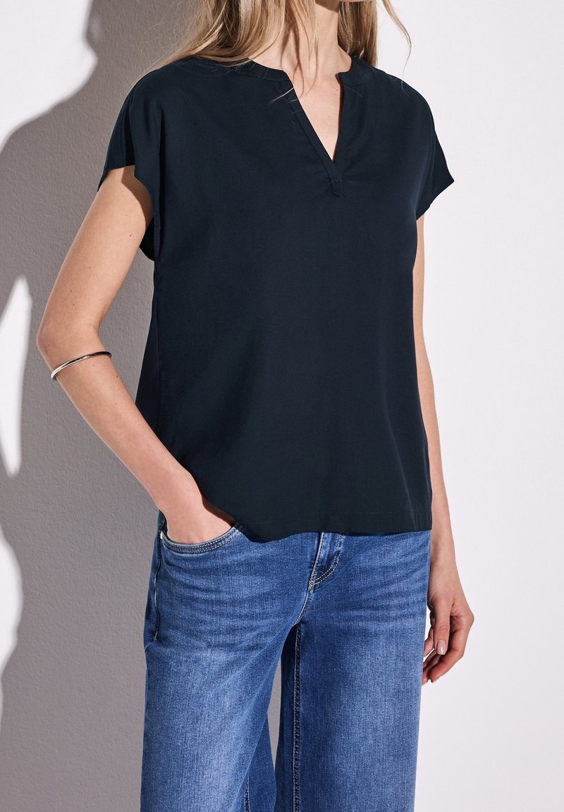 Femme portant un haut noir à manches courtes en V et un jean bleu, une main dans la poche, debout devant un mur uni.