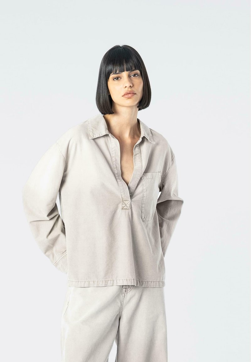 Camicia beige chiaro a maniche lunghe con colletto, una parziale abbottonatura e taschino frontale. Il tessuto è morbido e ha una vestibilità rilassata.