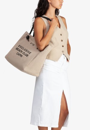 Borsa tote in tela beige con testo nero "EXCLUSIVE BEACH CLUB CAPRI" e tracolle nere regolabili, abbinata a un gilet chiaro e una gonna bianca.