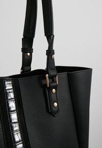 Sac fourre-tout noir en cuir synthétique texturé, bande d'accent argentée brillante et quincaillerie dorée. Doté de deux poignées pour le transport.