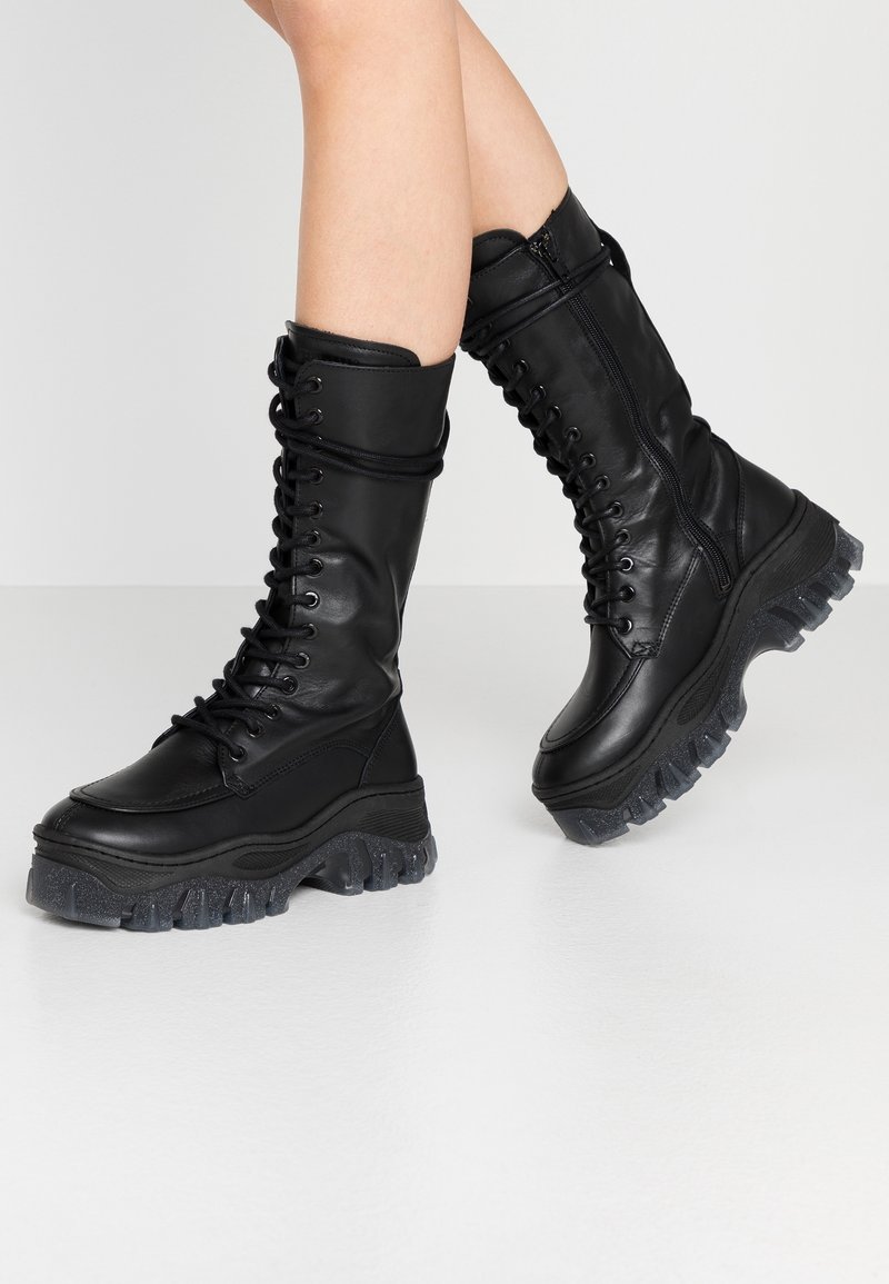 Bottes noires montantes jusqu'aux genoux en cuir lisse avec une semelle en caoutchouc épaisse et texturée, dotées d'un laçage à l'avant et d'une fermeture éclair latérale pour un port facile.