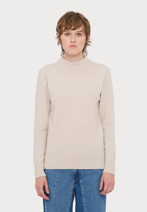 Lys beige genser med ribbestrikket høy hals, lange ermer og ribbestrikket kant. Myk tekstur, tettsittende silhuett, kombinert med blå denim.
