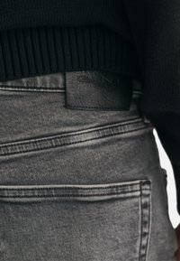 Szare dżinsy z denimu z czarną skórzaną naszywką z logo, z widocznymi detalami szwów oraz teksturowanym, gładkim wykończeniem.