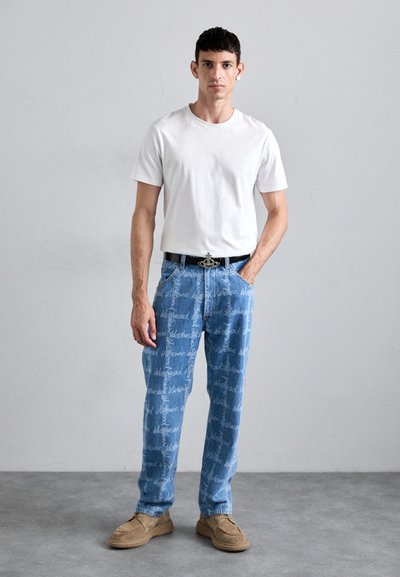 Cămașă pentru bărbați, compusă dintr-un tricou ajustat din bumbac alb, pantaloni din denim albastru cu un model repetitiv de logo și pantofi din suede maro.