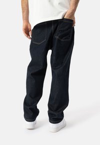 Temno denim hlače z udobnim krojem, z vidno rumeno šivanko in dvema zadnjima žepoma, v kombinaciji z belimi supergami.