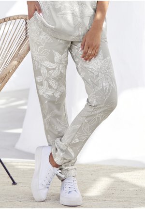 Lichtgrijze joggers met witte bloemenpatronen, elastische boorden en zijzakken, gecombineerd met witte sneakers op een gestructureerde ondergrond.