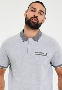 Grå polo med en kontrastfarvet sort og hvid kant langs kraven og ærmerne. Har en vandret lomme med stribet detaljering.