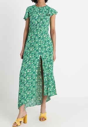 Vrouw draagt een groene midi-jurk met bloemenprint, een voorsplit en zwierige mouwen, gecombineerd met gele sandalen met hakken, staand tegen een effen achtergrond.