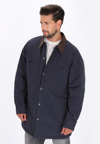 Mann mit Bart, der eine marineblaue knöpfbare Jacke mit braunem Kragen über einem weißen Hemd trägt und nach links schaut, vor einem schlichten weißen Hintergrund.