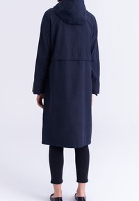 Manteau long bleu marine avec capuche, coupe ample et fente au dos. Confectionné dans un tissu lisse, associé à des leggings noirs et des chaussures à enfiler.