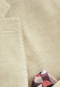 Tissu beige avec une texture fine, présentant un coin de poche cousu avec un mouchoir de poche orné de motifs dans des nuances de rose et de noir.