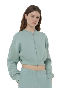 Sweatshirt verde chiaro corto con chiusura a zip, scollatura rotonda e spalle cadenti. Tessuto morbido con polsini e orlo a coste.