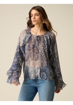 Donna che indossa una camicetta trasparente a maniche lunghe con motivi intricati blu e beige abbinata a jeans azzurri.