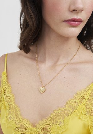 Kobieta w żółtej bluzce z koronkową lamówką i złotym naszyjnikiem z serduszkiem na cienkim łańcuszku, widoczna od ramion do dolnej części twarzy.