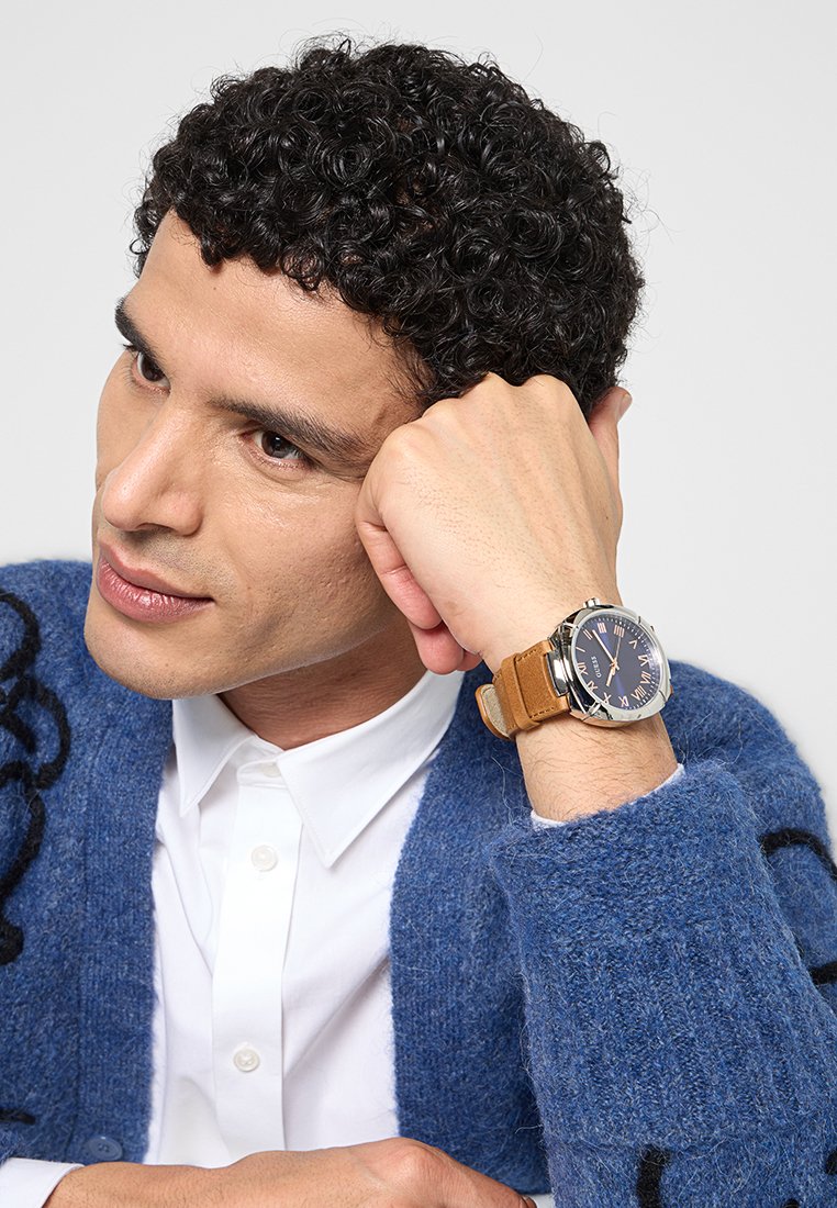 Reloj de tono plateado con esfera azul oscuro y correa de cuero color cuero natural. El modelo lleva un cárdigan azul con patrones negros y una camisa blanca.