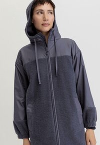 Giacca grigia con cappuccio, dotata di un pannello frontale in morbido pile, maniche in tessuto liscio e tasche ai fianchi. Coulisse regolabile sul cappuccio.